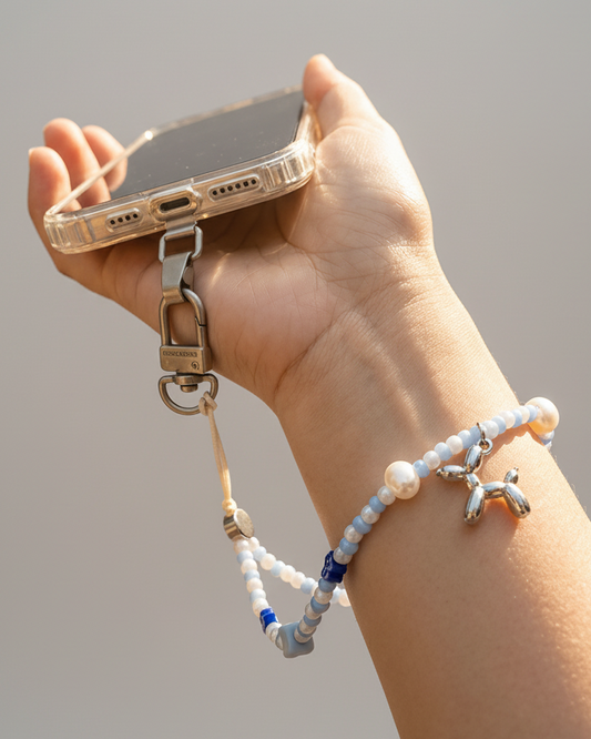 Hand holding a phone with a beaded strap, outdoor setting with blurred background