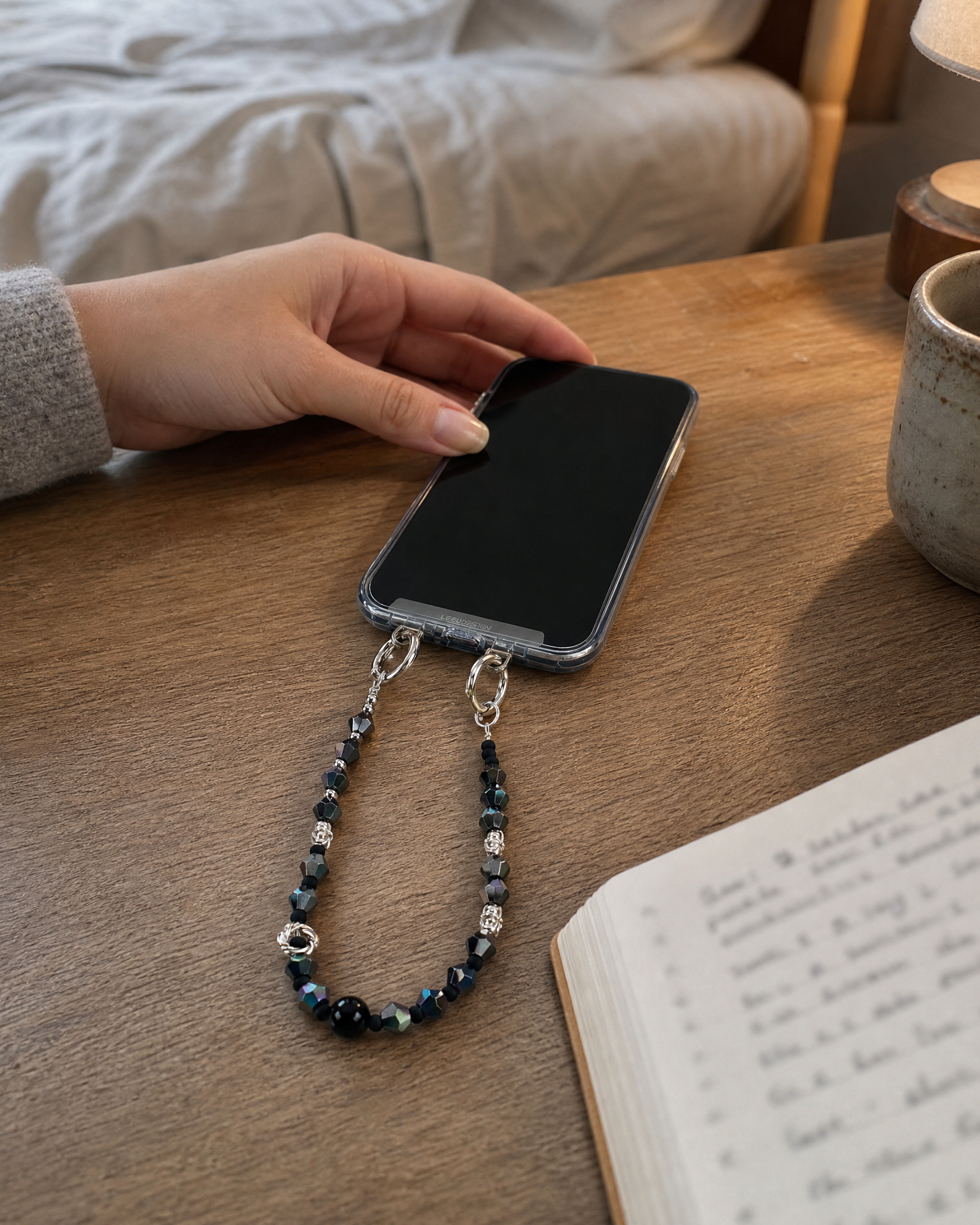 A close-up lifestyle shot of a person's hand holding a smartphone with a dark blue beaded wrist strap attached to the bottom. The scene is set on a wooden table with a book, a ceramic mug, and a lamp in the background.