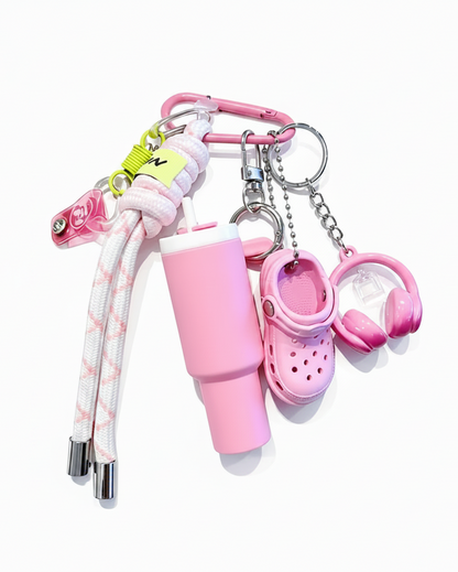 A pink-themed keychain bag set featuring a mini pink tumbler, a croc-heel keychain, pink headphones, a braided pink-and-white strap, and a pink carabiner on a white background.