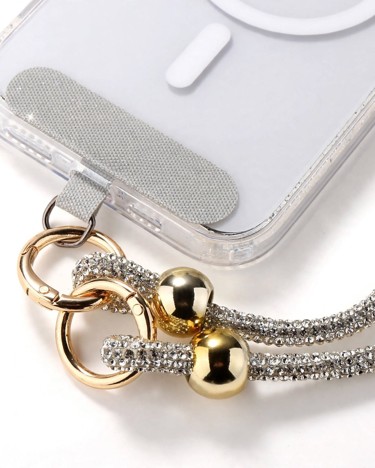 A close-up of a clear phone case with a decorative wrist strap featuring a gold-tone ring clasp, two shiny gold beads, and silver rhinestone-covered straps on a white background.
