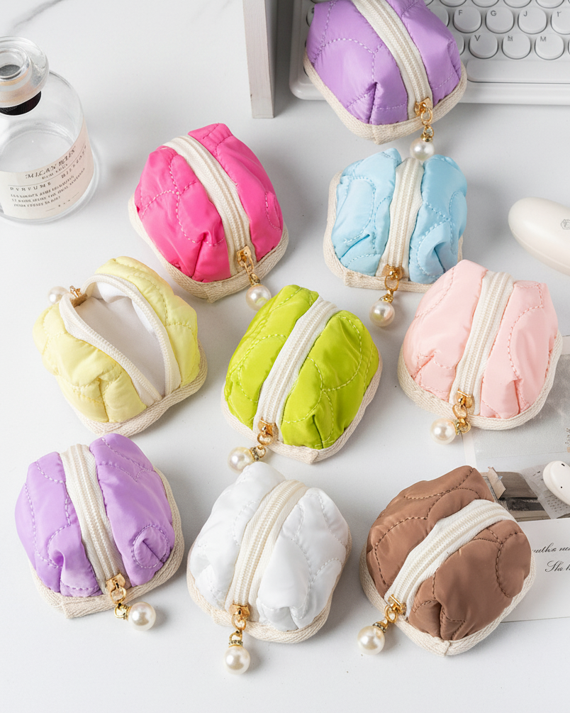 Nine small quilted zippered coin pouches in assorted pastel and neutral colors, arranged on a white desk with a keyboard, bottle, and other desk props visible.